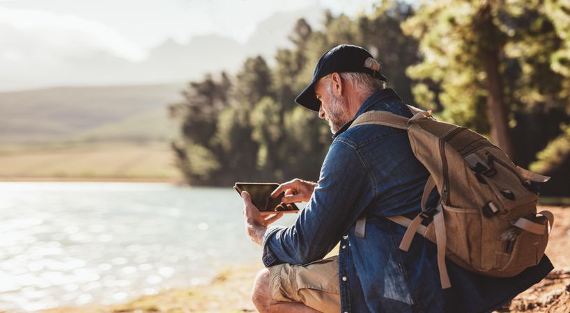 Mature man with backpack using digital tablet while sitting near a lake