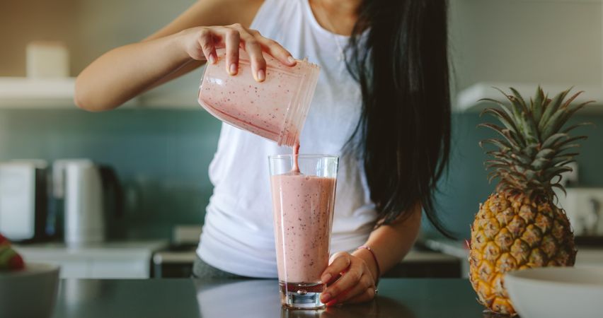Woman pouring healthy smoothie in glass from grinder jar on kitchen counter