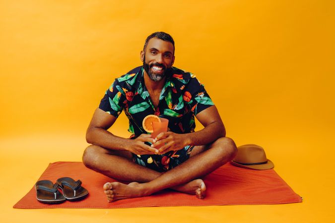 Black male sitting on towel and smiling at camera while holding tropical drink