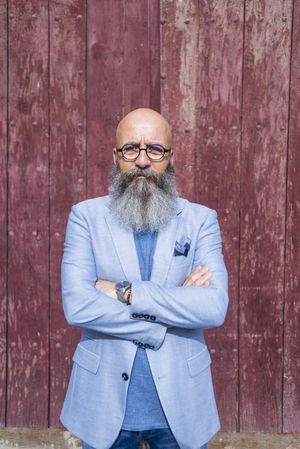 Front view of a mature trendy bearded hipster standing against a vintage red door outdoors while looking to camera