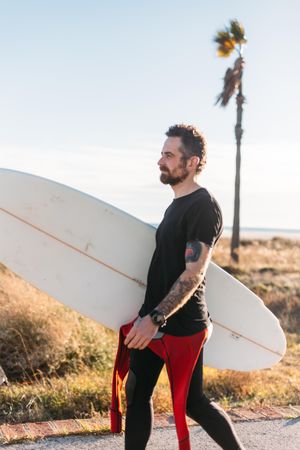 Tattooed man standing on road near the ocean holding surfboard