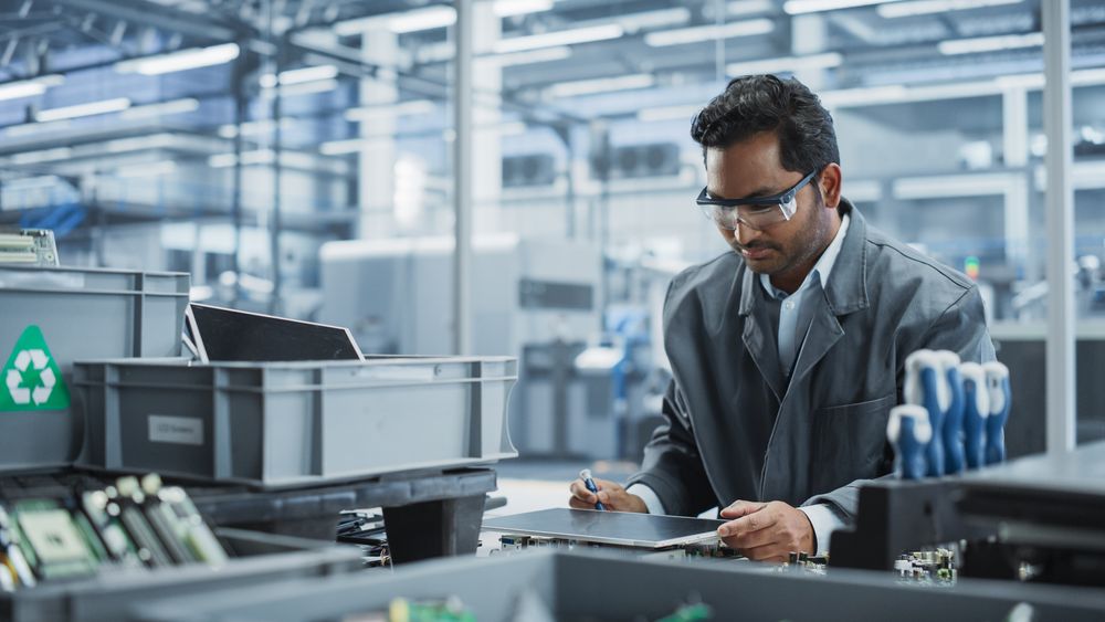 Indian Male Worker Disassembling Laptops To Recycle Electronic Components For Microchip Production At Automated Electronics Factory With Robotic Arms.