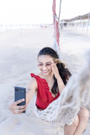 A young Hispanic woman is smiling taking a selfie photo on a hammock at the beach
