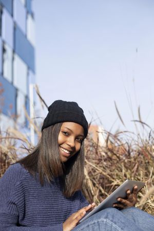 Female in hat and sweater sitting outside with tablet, copy space