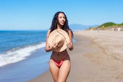 A woman in a stylish bikini strolls along the beach, wearing a straw hat and enjoying the sun
