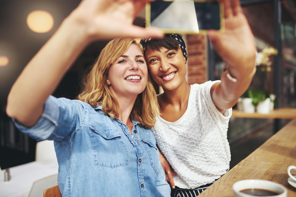 Two female friends sitting at a cafe table smiling and taking a selfie
