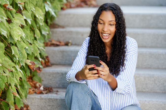 A happy young woman engaged with her smartphone while sitting on steps among greenery