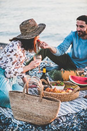 Couple having picnic by the ocean with man holding strawberry as woman takes a bite