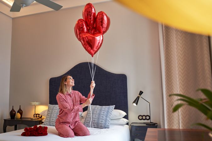 Happy female sitting on bed with red heart balloons