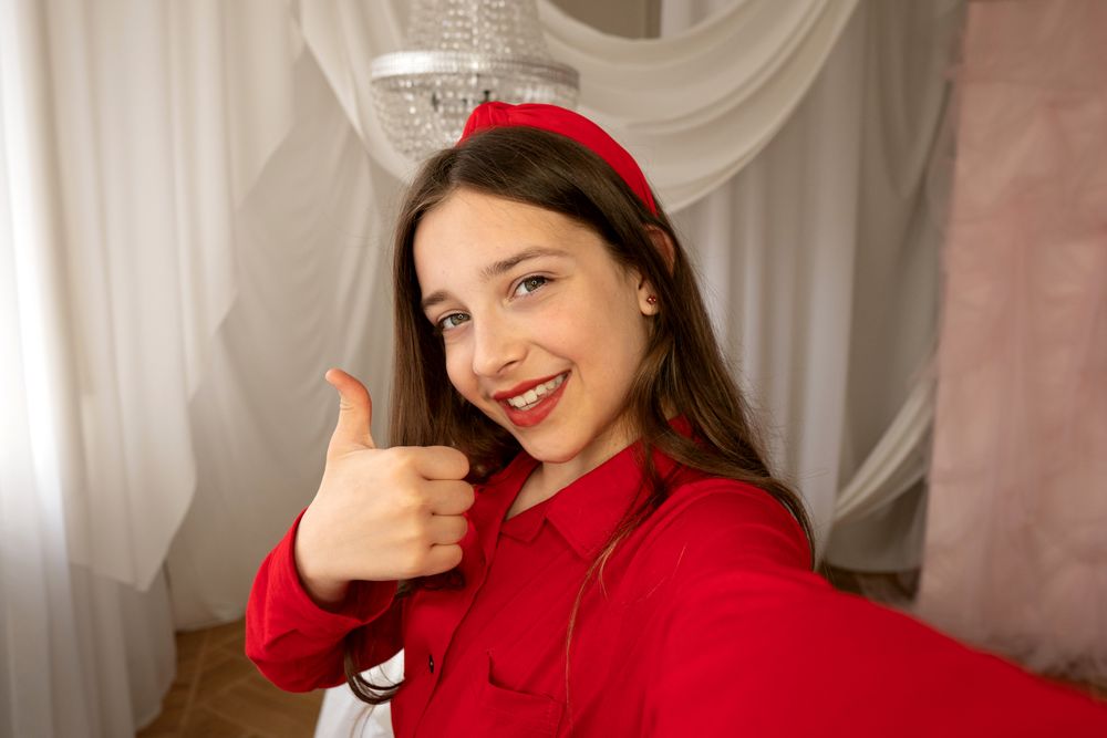 Teen girl smiling giving thumbs-up gesture in selfie photo.