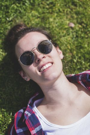 Above view of a young smiling hipster woman lying on grass in a sunny day at a park while taking a selfie with a mobile phone