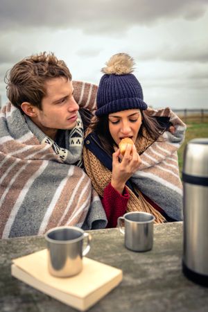 Cute couple wrapped in blanket on cold park bench with coffee flask and muffin, vertical