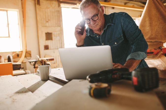 Middle aged carpenter working on laptop and answering phone call in his workshop