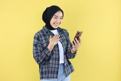 Happy Excited Asian Muslim Woman Wearing Hijab Holding Smartphone while TOuch Her Chest Smiling Cheerfully, Isolated on Yellow Background.
