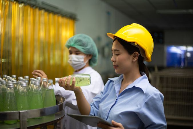 Asian female manager and worker holgin digital tablet checking information and quality control for juice drink product on bottling factory machinery.