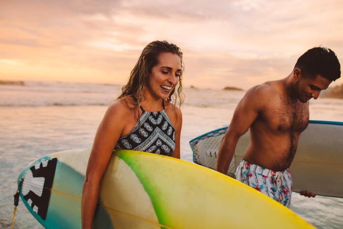 Smiling man and woman walking on the beach carrying surfboards