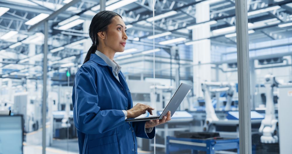Electronics Manufacturing Technician Working on a Laptop Computer at a Factory. Asian Female Specialist Developing, Maintaining, Setting Up Programs for Machine Production