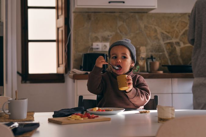 Cute boy eating from spoon and cup in kitchen