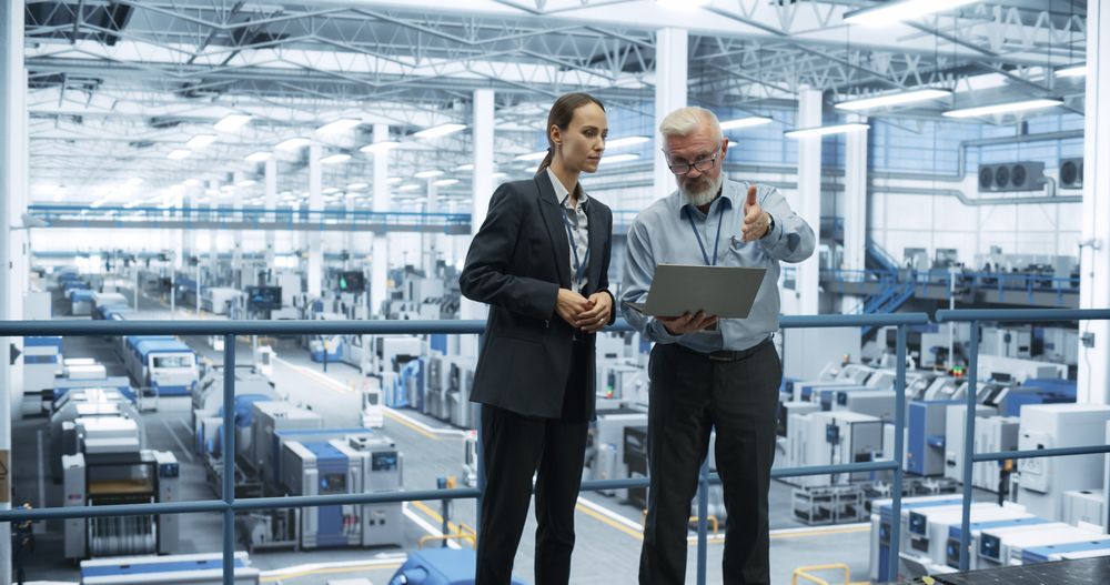 Male and Female Engineers Standing at a Plant, Using Laptop Computer and Talking in a Factory Facility with Equipment Producing Modern Electronic Components for Different Industries