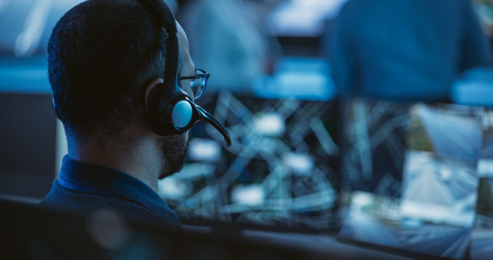 Technical Customer Support Specialist is Talking on a Headset while Working on a Computer in a Call Center Control Room Filled Display Screens and Data Servers.