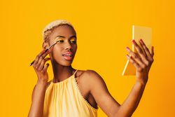 Black woman with short blonde hair holding a pocket mirror applying gold eyeshadow