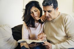 couple relaxing on sofa reading a book together and pointing at text with curiosity