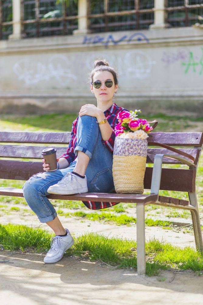Front view of a young hipster woman sitting on a park bench relaxing in a sunny day while looking to camera