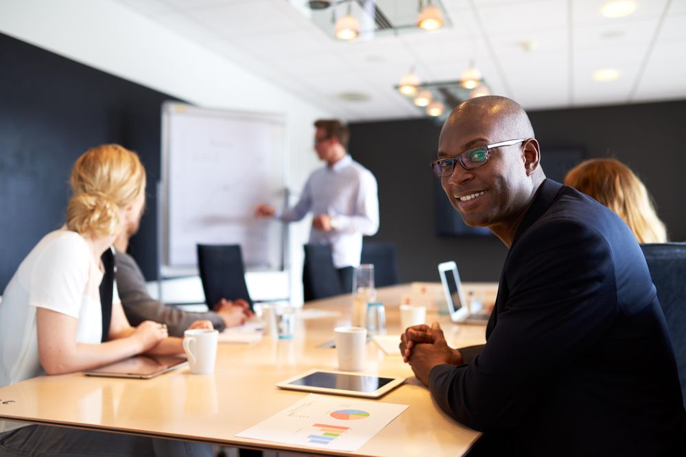 Businessman smiling at camera while his colleague gives presentation in the background
