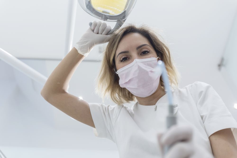 Dentist wearing surgical mask while holding angled mirror and drill, ready to begin