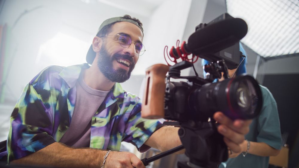 Young Male Cinematographer With Beard And Glasses, Smiling While Operating A Professional Camera In A Studio Setting, Capturing Footage For A Creative Advertising Project.