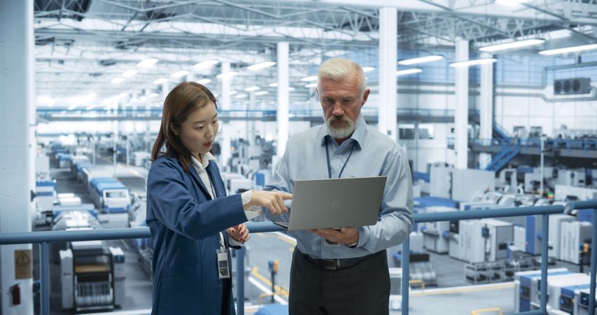 Male Engineer and Female Project Manager Standing on a Platform, Talking, Using Laptop Computer and Monitoring Production at a Modern Automated Electronics Manufacture with AI Robots