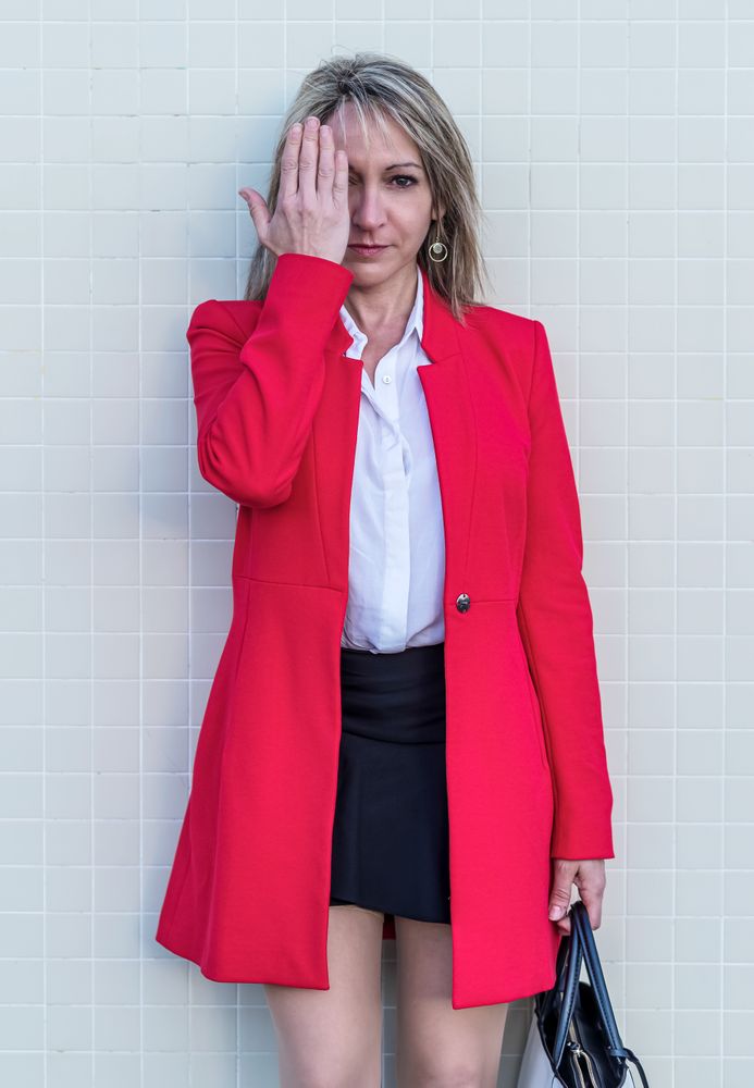 Portrait of a blonde woman wearing red jacket and skirt against a tiled wall covering her eye