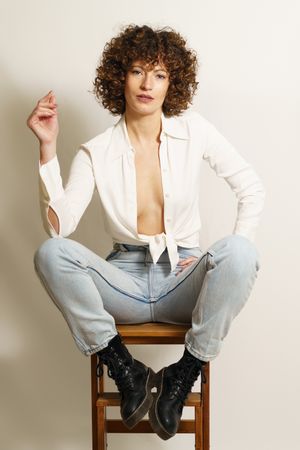 Self-assured woman in jeans and boots sitting on wooden stool in studio shot looking at camera