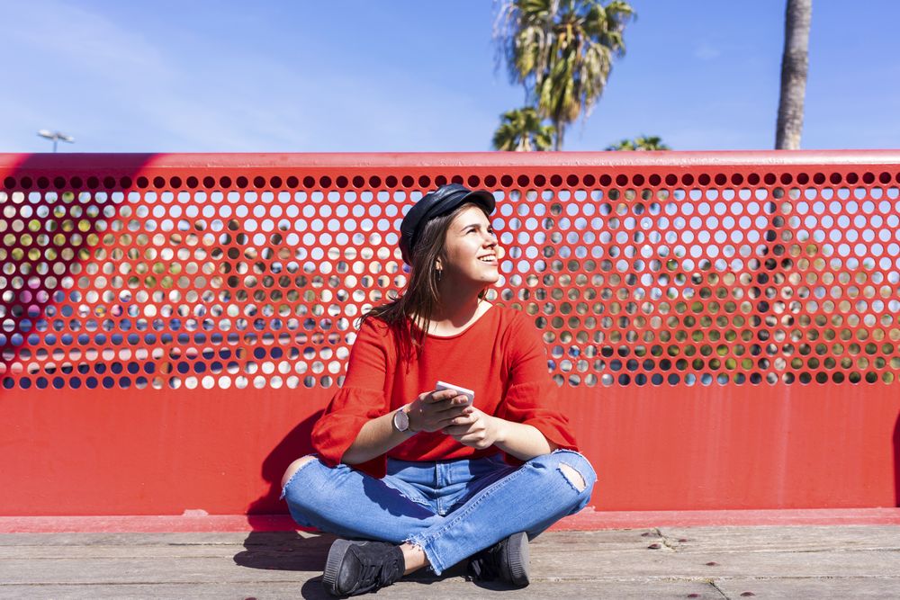 Happy woman in red sitting cross legged using a mobile phone outdoors on a bright day