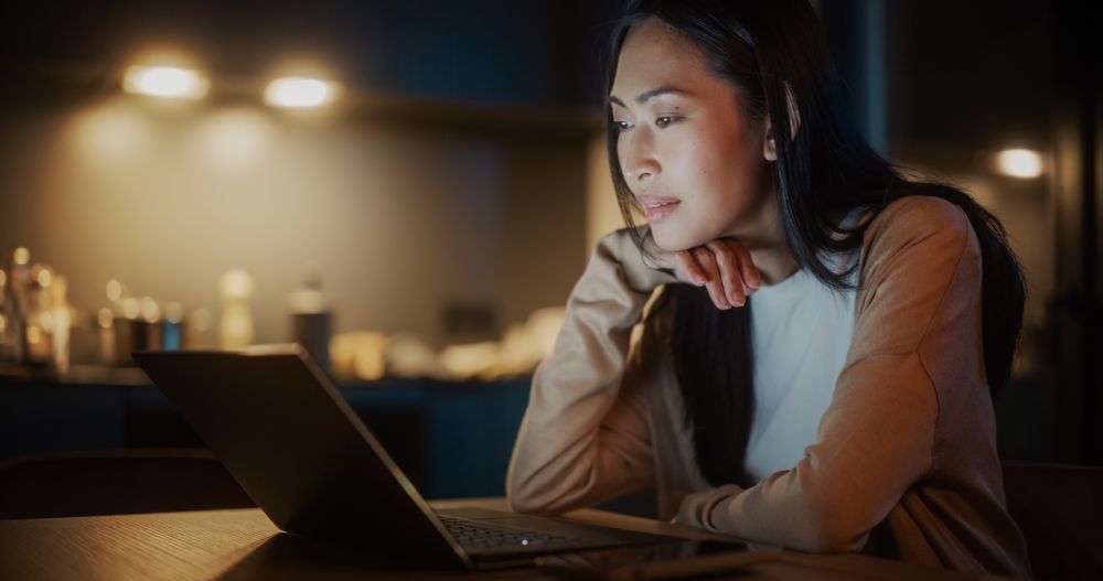 Authentic Asian Woman Sitting at a Table in a Cozy Kitchen and Using Laptop Computer at Home at Night.