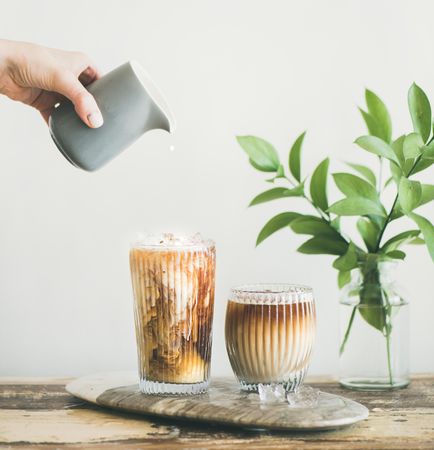 Hand dropping cream into iced coffee, square crop with leaves in background