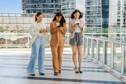 Three businesswomen are walking while using smartphones outdoors. All women are dressed in business attire..