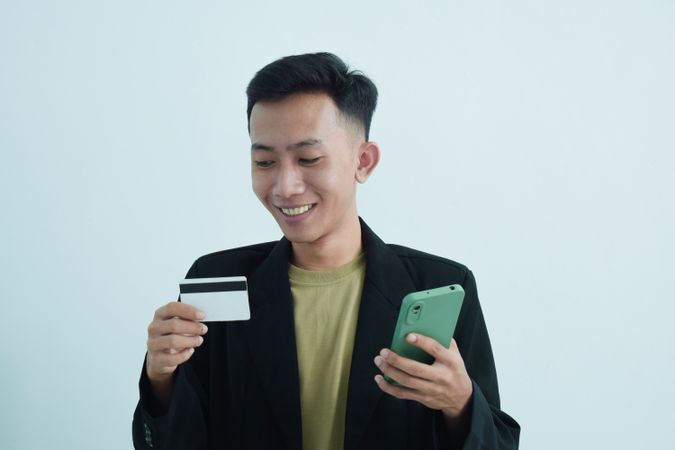Smiling Asian businessman in suit holding blank credit card and smartphone