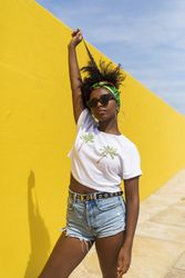 Young Black woman with sunglasses standing against yellow wall and playing with hair