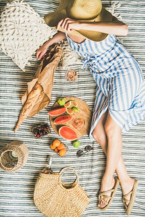 Woman lounging on picnic blanket with thatched bag with glass of rose, baguette, sliced fruit