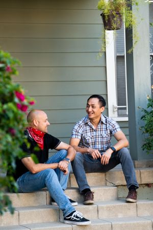 Two men laughing and smiling sitting on the steps in the front of a green house with plants around