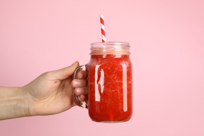 Female hand holds glass jar of strawberry cocktail on pink background, close up