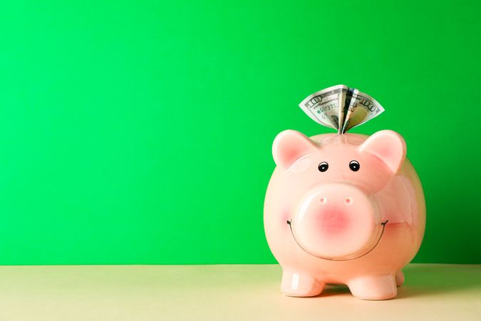Happy piggy bank with one hundred dollar on color table against green background, space for text. Finance, saving money