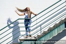 Joyful female in denim overalls standing on skateboard on stairs playing with her hair 5nXEMb