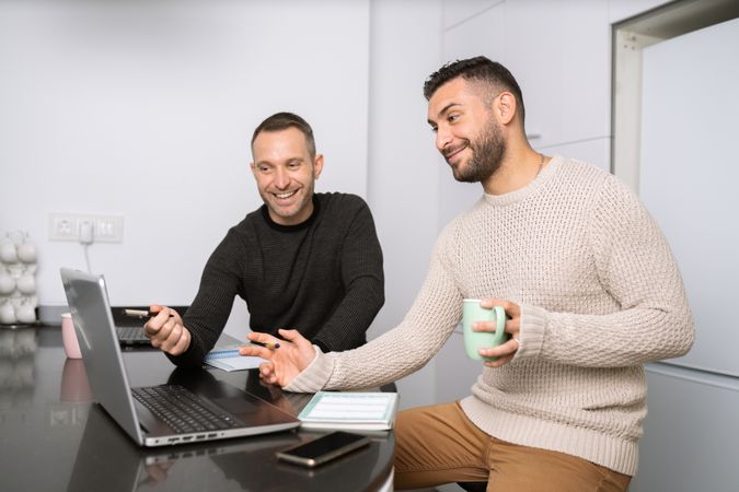 Two men sitting at kitchen counter entertained by laptop screen