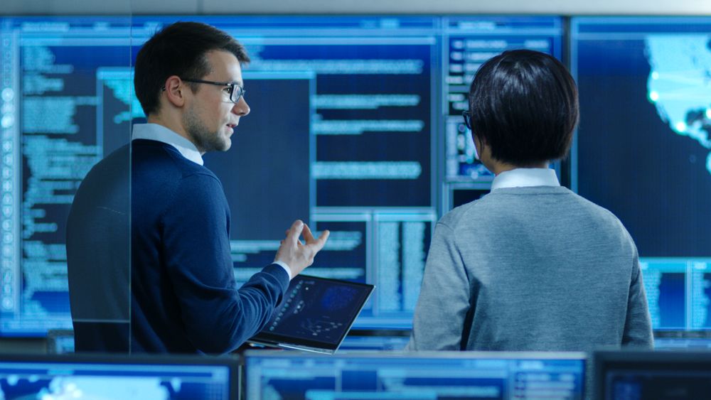In the System Control Room IT Specialist and Project Engineer Have Discussion while Holding Laptop, they're surrounded by Multiple Monitors with Graphics.