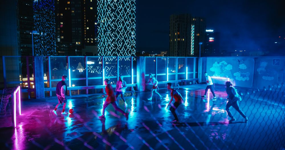 Group of Diverse People Playing Soccer at a Stylish Rooftop Spot with Neon Lights and Skyscrapers at Night.