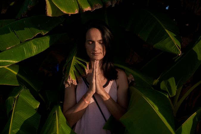 Peaceful Hispanic woman with prayer hands near green trees