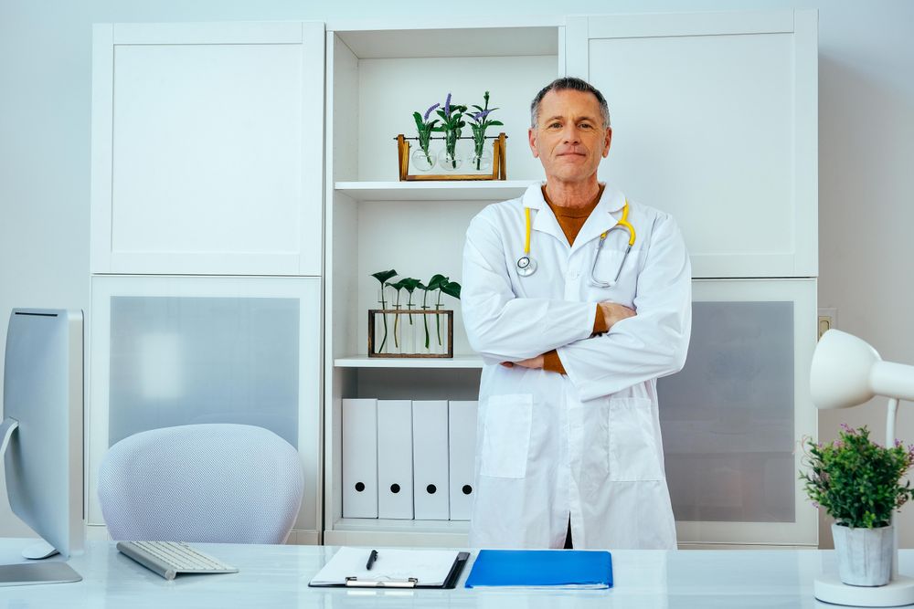 Mature male doctor standing in his office with arms crossed