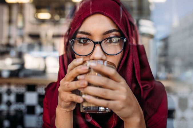 Close up portrait of young woman wearing hijab and eyeglasses drinking coffee